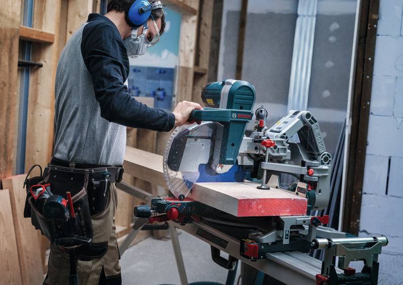 Eine Person in Schutzausrüstung nutzt eine Elektrosäge, um Holz in einer Werkstatt zu schneiden, umgeben von Werkzeugen und Baumaterialien.