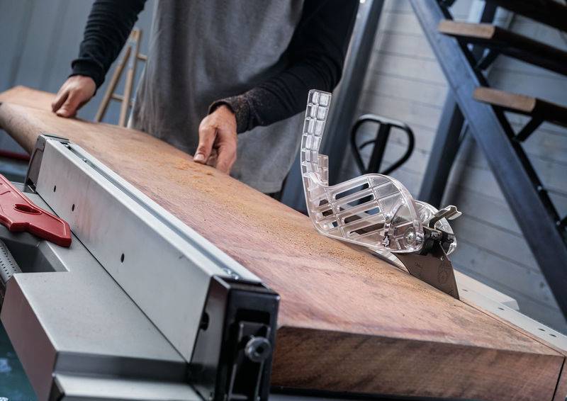 Eine Person, die ein Holzbrett mit einer Tischkreissäge in einer Werkstatt zuschneidet, wobei der Fokus auf Präzision und Sicherheit liegt, mit einem Schiebestock in der Nähe.