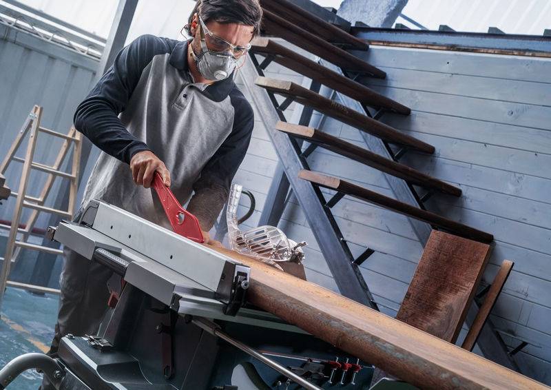 Ein Mann in Schutzausrüstung benutzt eine Tischkreissäge, um Holz in einer Werkstatt mit Treppen und verschiedenen Werkzeugen im Hintergrund zu schneiden.