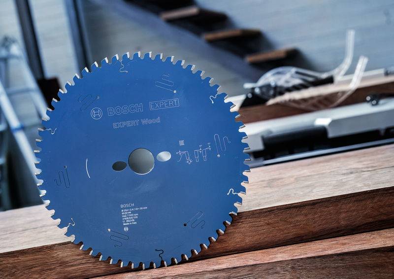 Ein Bosch Expert Holz-Kreissägeblatt lehnt an Holzbrettern, mit Treppen im Hintergrund, was auf eine Schreinerei hindeutet.