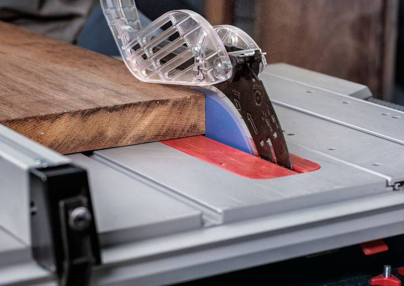 Eine Tischkreissäge schneidet ein Stück Holz, mit einer klaren Schutzvorrichtung über dem Sägeblatt, in einer Werkstattumgebung.