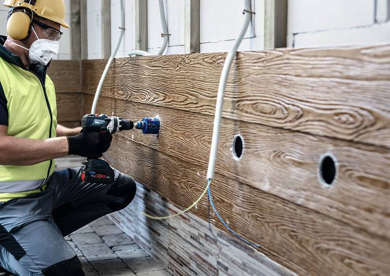 Ein Arbeiter mit Schutzhelm und Maske benutzt einen Elektroschrauber, um ein Loch in eine Holzwand zu bohren, umgeben von teilweise gebohrten Löchern.