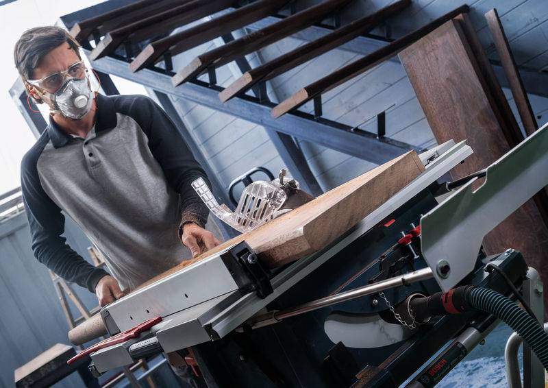 Eine Person in Schutzausrüstung bedient eine Tischkreissäge, um ein Stück Holz in einer Werkstattsetting mit Regalen im Hintergrund zu schneiden.