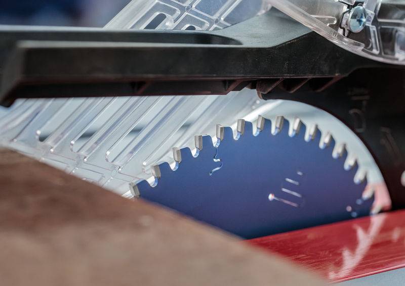 Eine Nahaufnahme eines Kreissägeblatts hinter einer durchsichtigen Kunststoffschutzvorrichtung, montiert auf einem roten Tisch, die seine scharfen metallischen Zähne hervorhebt.