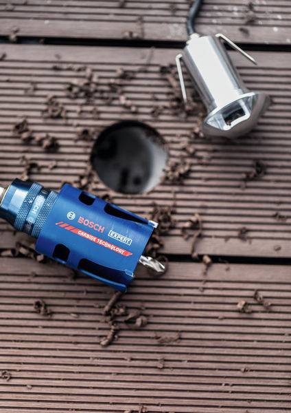 Ein blaues Bosch-Elektrowerkzeug und eine metallische Leuchte auf einer Holzoberfläche mit einem Loch und verstreuten Holzspänen.