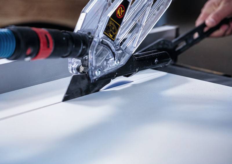 „Eine Säge, die ein Stück Holz schneidet, mit einer Schutzvorrichtung und einem blauen Absaugschlauch ausgestattet, bedient von einer Person.