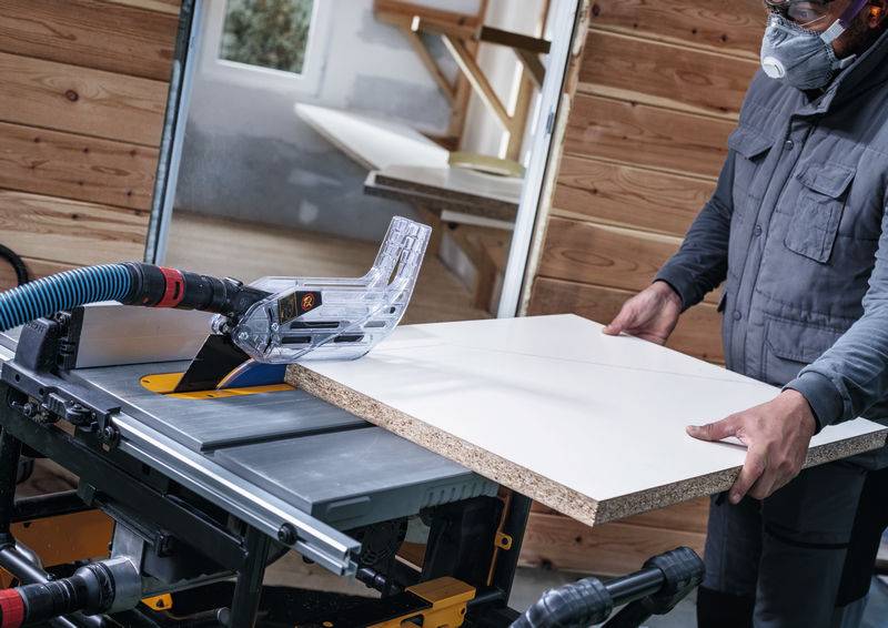 Eine Person in Schutzausrüstung bedient eine Tischkreissäge und schneidet ein Holzbrett in einer Werkstatt.