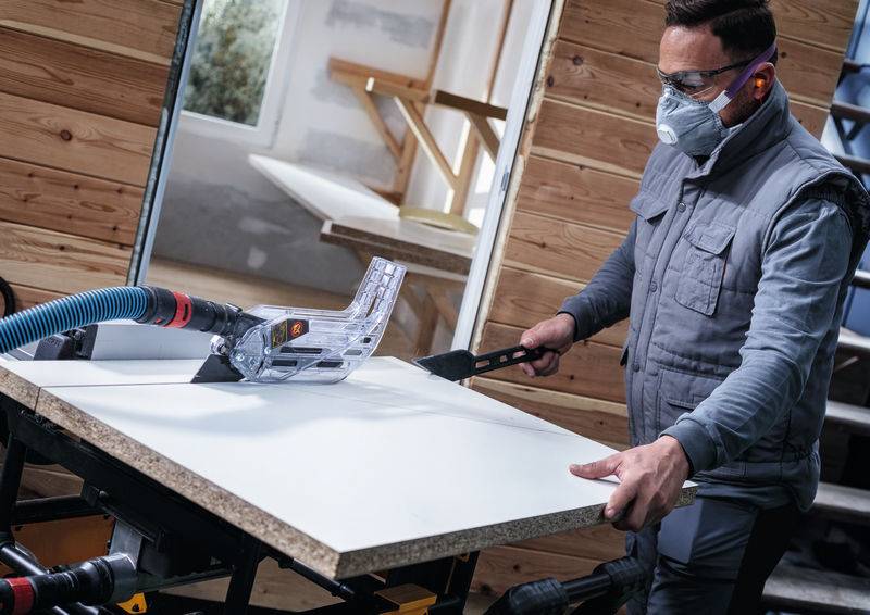 Eine Person in Schutzausrüstung verwendet ein Holzbearbeitungswerkzeug, um ein großes Stück weißes Material auf einer Werkbank in einer Werkstatt zu schneiden.