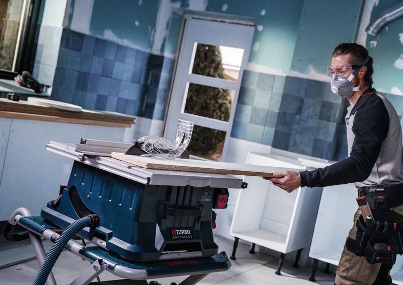 Eine Person in Schutzausrüstung benutzt eine Tischkreissäge, um Holz in einem teilweise renovierten Raum mit blauen Wänden und Bauwerkzeugen zu schneiden.