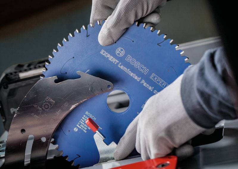 Handschuhe tragende Hände halten ein blaues Bosch-Kreissägeblatt mit gekennzeichneten Merkmalen, positioniert in der Nähe eines Schneidwerkzeugs, das Präzisionstechnik zeigt.