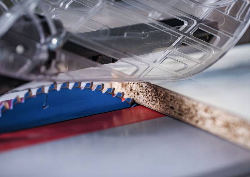 Eine Kreissägeblatt schneidet durch ein Stück Sperrholz, wobei das Schutzgehäuse sichtbar ist. Die Szene betont industrielle Holzbearbeitung.