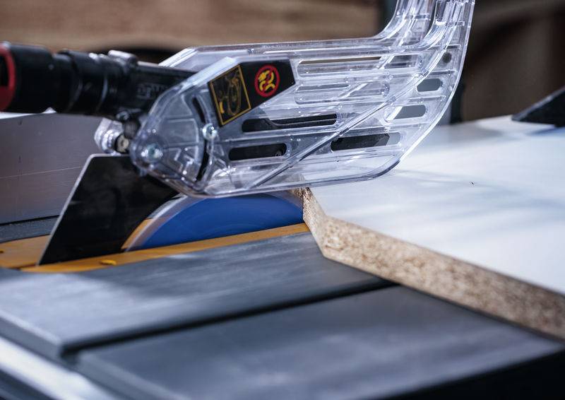 Eine Tischkreissäge schneidet ein Stück Holz, mit einer Schutzvorrichtung über dem rotierenden Sägeblatt, was die Verwendung von Holzbearbeitungsausrüstung veranschaulicht.