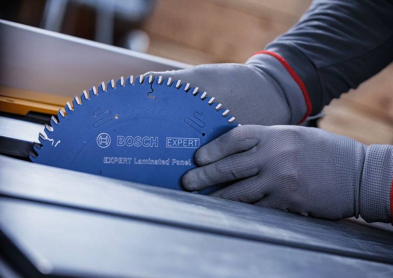 Person trägt Handschuhe und handhabt ein Bosch Expert-Sägeblatt an einer Tischkreissäge, wobei der Fokus auf Sicherheit und präzisem Schneiden in einer Werkstattumgebung liegt.