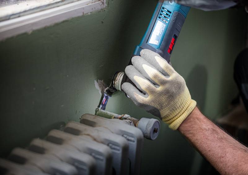 Eine Person nutzt ein Elektrowerkzeug, um ein Rohr zu schneiden oder zu schleifen, das mit einer Heizung neben einer Wand verbunden ist. Sie trägt Handschuhe zum Schutz.