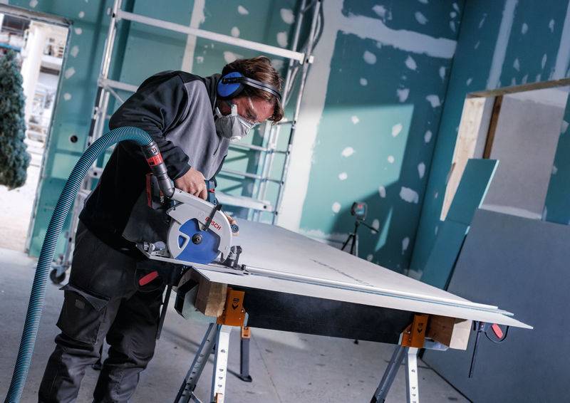 Eine Person in Schutzausrüstung schneidet mit einer Kreissäge ein großes Paneel auf einer Baustelle zu. Verstreute Werkzeuge und Baumaterialien sind sichtbar.
