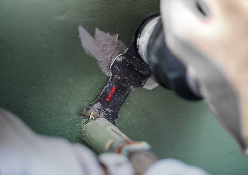 Eine Person nutzt ein Elektrowerkzeug, um ein Rohr gegen eine grüne Oberfläche zu schneiden; das Etikett des Werkzeugs trägt die Aufschrift 