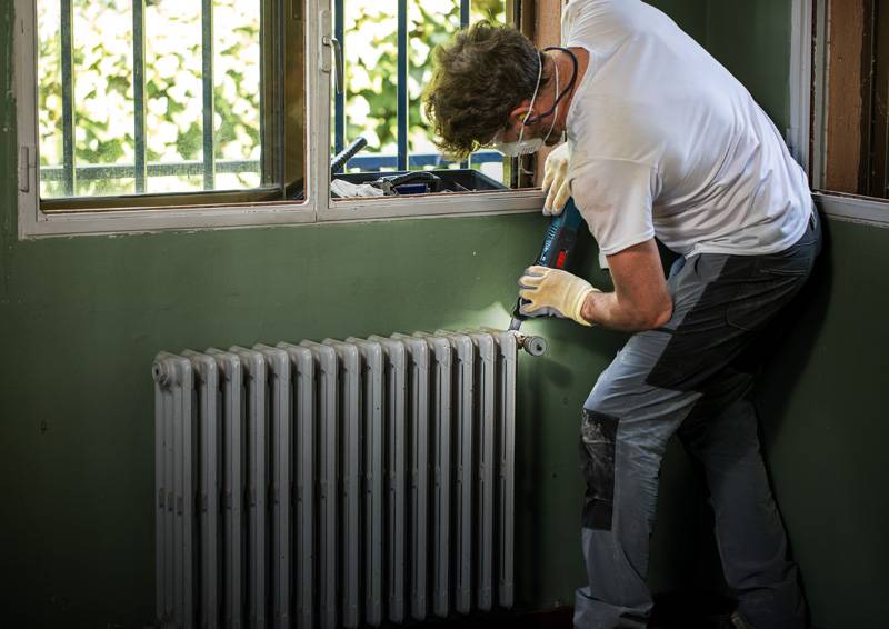 Eine Person in Schutzausrüstung bohrt in die Wand über einem Heizkörper in einem Zimmer mit grünen Wänden und vergitterten Fenstern.
