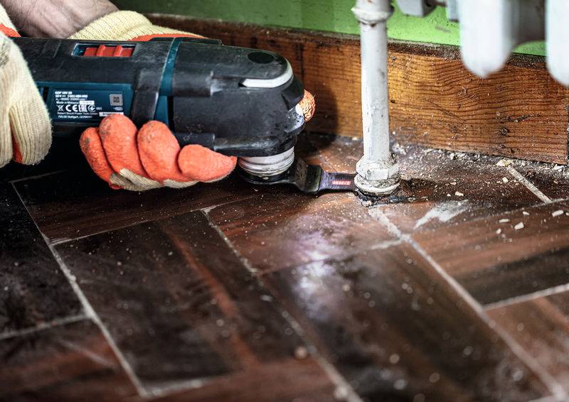 Eine Person mit Handschuhen benutzt ein oszillierendes Werkzeug, um ein Rohr neben einem Holzboden und einer grünen Wand zu schneiden, was auf eine Renovierungsaufgabe hindeutet.
