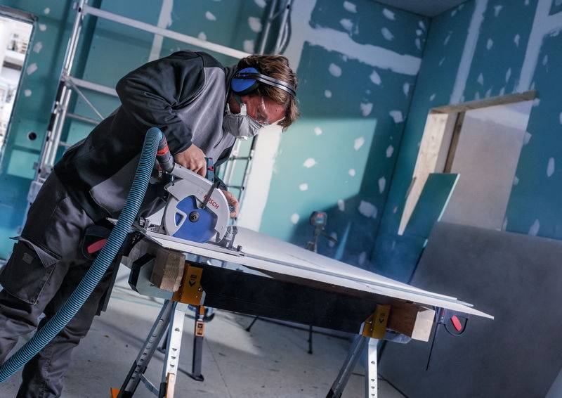 Eine Person in Schutzausrüstung schneidet ein großes Brett mit einer Elektrosäge auf einer Werkbank in einem teilweise konstruierten Raum.