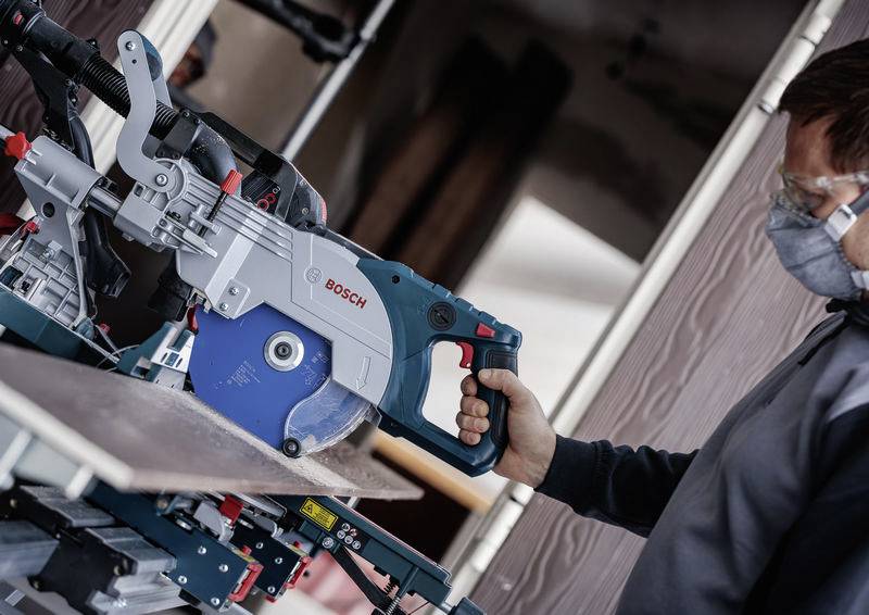 Eine Person mit Schutzbrille bedient ein Bosch-Elektrowerkzeug und schneidet ein Stück Holz in einer Werkstattumgebung.