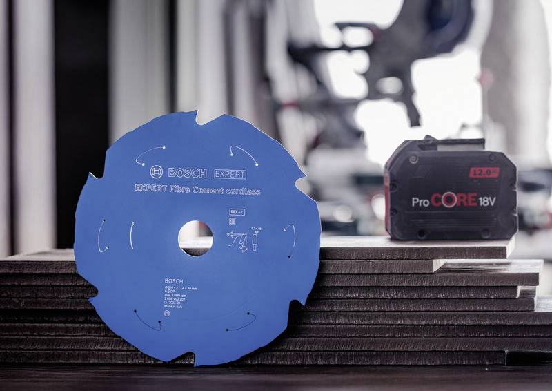 Ein blaues Bosch Expert Kreissägeblatt für Faserzement wird auf gestapelten Platten neben einem ProCORE 18V Akku in einer Werkstatt angezeigt.