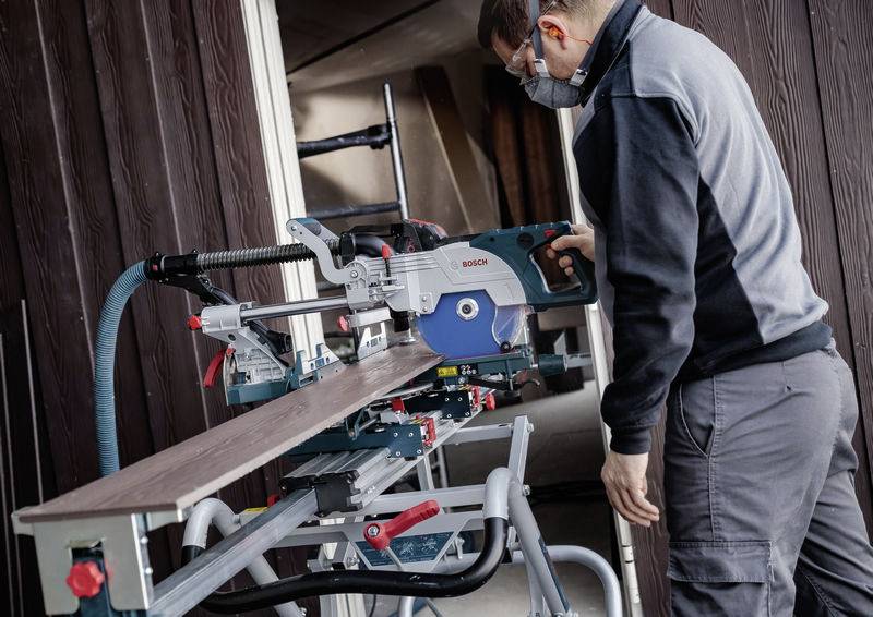 Eine Person bedient eine Elektrosäge und schneidet ein langes Holzbrett in einer Werkstattumgebung. Sie trägt Schutzausrüstung, einschließlich einer Maske und eines Gehörschutzes.