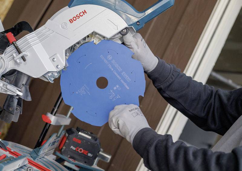 Eine Person, die Handschuhe trägt, installiert ein blaues Sägeblatt in eine Bosch-Gehrungssäge. Der Hintergrund zeigt einen Teil einer Holzkonstruktion.