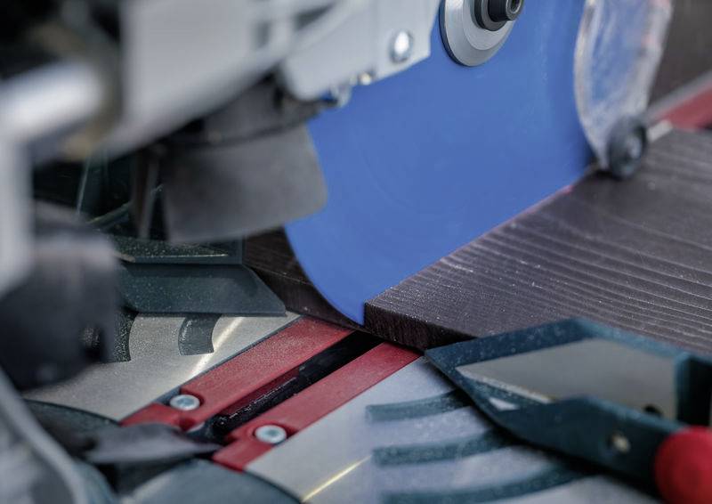 Eine Nahaufnahme einer Kreissäge, die ein Stück dunkles Holz schneidet. Das Sägeblatt ist blau, und das Holz ist an roten und silbernen Führungen ausgerichtet.