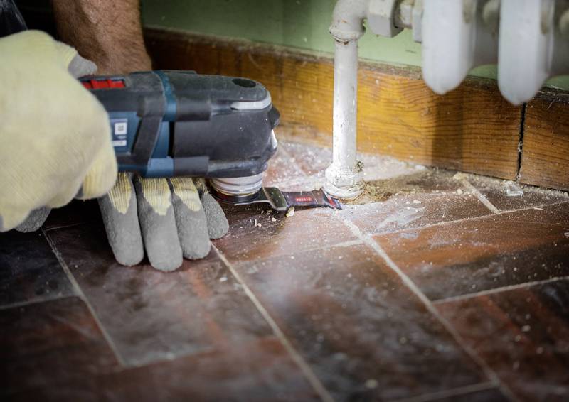 Eine Person nutzt ein Elektroschleifergerät, um eine Holzleiste in der Nähe eines Heizkörperrohrs durchzuschneiden, was laufende Heimwerkerarbeiten zeigt.