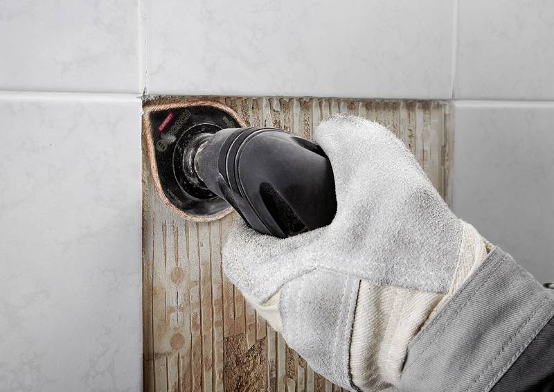 Eine behandschuhte Hand hält ein Elektrowerkzeug gegen eine Badezimmerfliese und reinigt Fuglinien. Die Fliese ist teilweise gereinigt und zeigt einen Vorher-Nachher-Effekt.