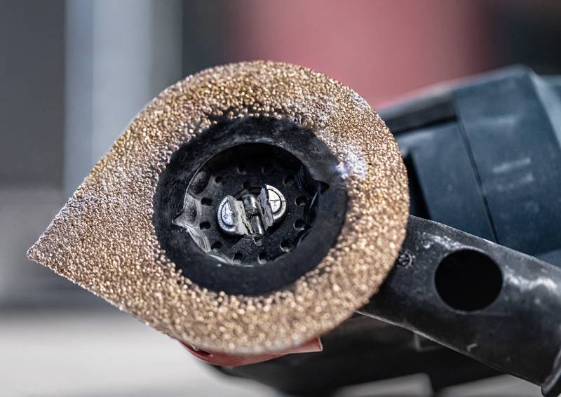 Eine Person benutzt einen Elektroschrauber mit einer Lochsäge, um ein Bohrloch in einen Holzblock zu bohren und dabei Sägespäne auf einer Betonoberfläche zu erzeugen.