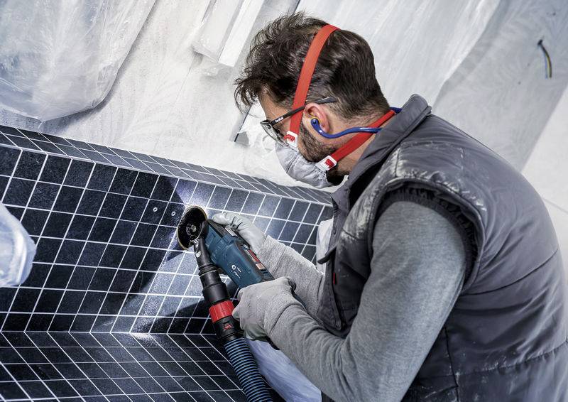 Ein Mann in Schutzausrüstung nutzt ein Elektrowerkzeug, um schwarze Fliesen in einer Bau- oder Renovierungsumgebung zu schneiden.