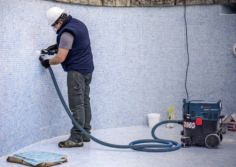 Ein Arbeiter, der ein Elektrowerkzeug auf einer gefliesten Oberfläche in einem leeren Schwimmbecken verwendet, trägt Schutzausrüstung und hat einen blauen Staubsauger neben sich.