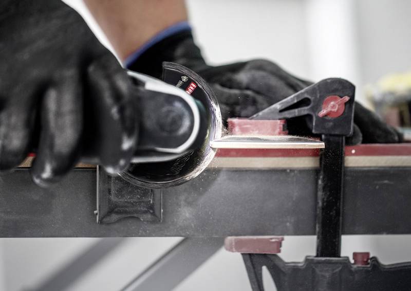 Eine Person mit schwarzen Handschuhen verwendet ein Elektrowerkzeug, um die Kante eines Skis zu trimmen, der an einer Werkbank eingespannt ist.