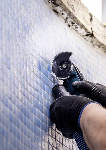 Eine Person verwendet einen Winkelschleiffer zum Schneiden oder Polieren von blauen Fliesen an einer Wand, trägt schwarze Handschuhe und zeigt einen Fliesenerneuerungsprozess.