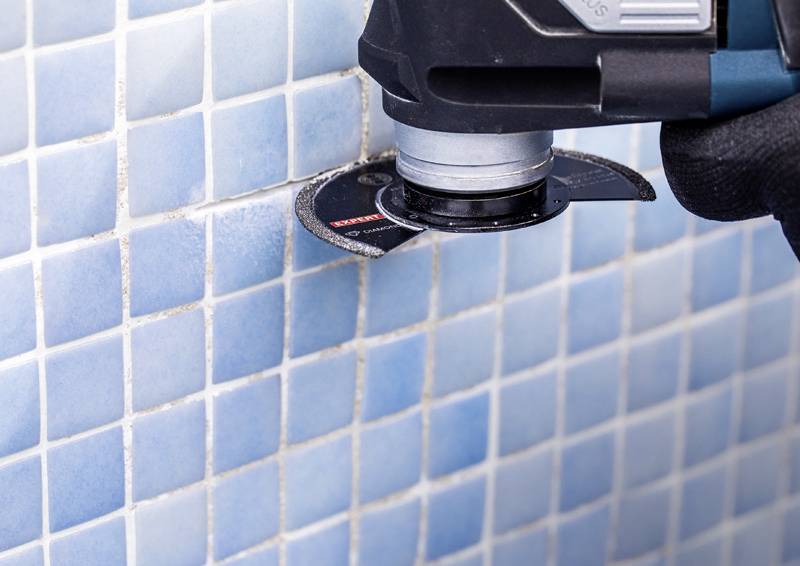 Ein Elektrowerkzeug mit einer rotierenden Klinge wird verwendet, um Mörtel zwischen blauen und weißen Fliesen an einer Wand zu schneiden.