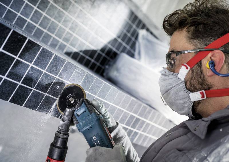 Ein Arbeiter in Schutzausrüstung verwendet ein Elektrowerkzeug, um eine geflieste Oberfläche zu polieren, wobei er sich auf einen bestimmten Bereich konzentriert.