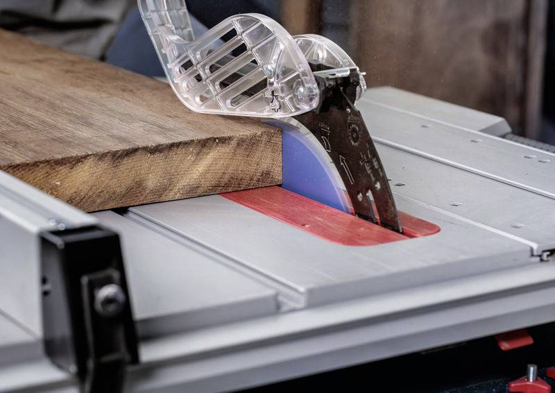 Ein Tischkreissäge mit Schutzvorrichtung schneidet ein Stück Holz. Das Sägeblatt ist teilweise angehoben und bereit zum Schneiden. Sicherheitsmerkmale sind sichtbar.