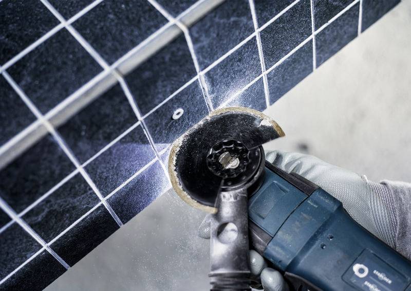 Ein Elektrowerkzeug schneidet durch dunkle Fliesen mit weißen Gitternetzlinien. Staub ist vom Schneideprozess sichtbar. Eine behandschuhte Hand hält das Werkzeug.