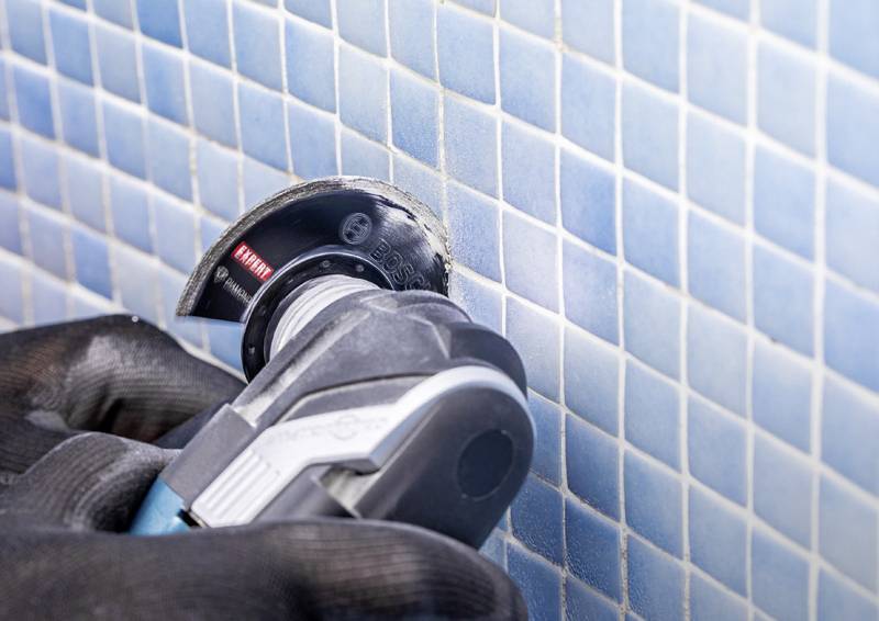 Eine behandschuhte Hand nutzt ein Elektroschleifergerät, um blaue Fliesen an einer Wand zu schneiden, präzise und vorsichtige Arbeit für eine Renovierungs- oder Reparaturaufgabe.
