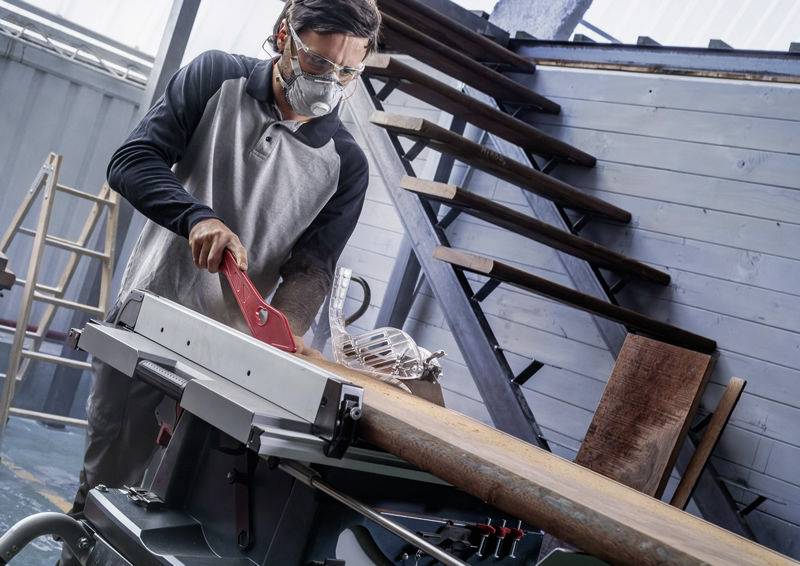 Eine Person in Schutzausrüstung bedient eine Tischkreissäge in einer Werkstatt und schneidet ein Holzbrett zu. Im Hintergrund sind Treppen und Werkzeuge sichtbar.