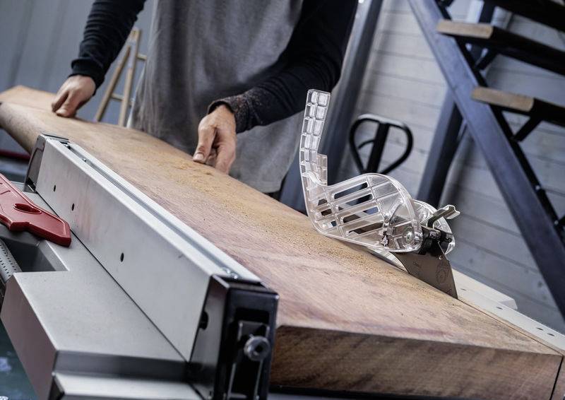 Eine Person bedient eine Tischkreissäge und schneidet ein großes Holzbrett. Eine Sicherheitsvorrichtung ist sichtbar, und der Arbeitsbereich umfasst metallene Treppen im Hintergrund.