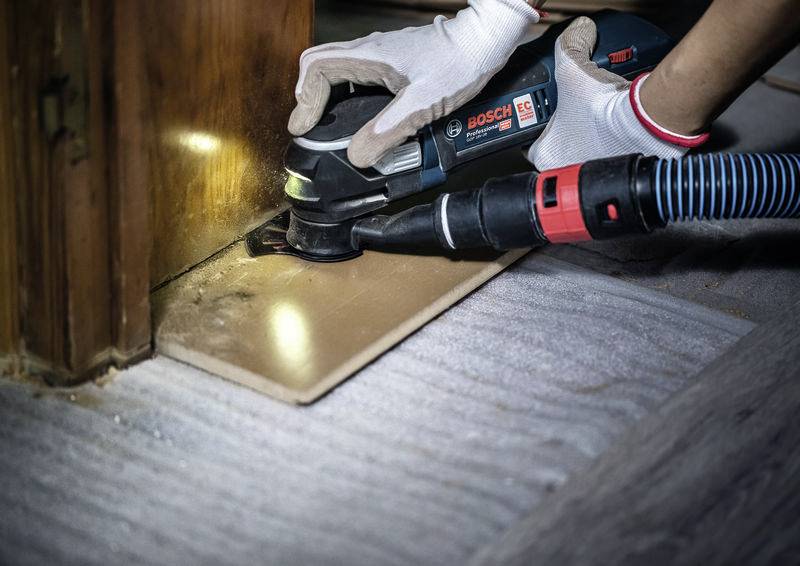 Person, die ein Bosch-Elektrowerkzeug zum Schleifen einer Holzoberfläche in der Nähe einer Wand verwendet, trägt weiße Handschuhe zum Schutz.