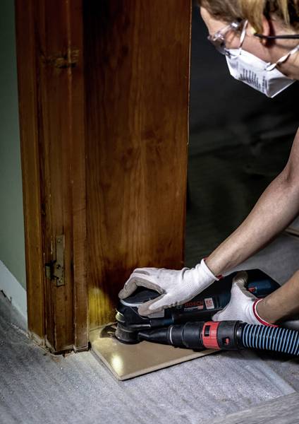 Eine Person mit Handschuhen und einer Maske benutzt ein Elektrowerkzeug, um eine Fliese am unteren Rand eines Holztürrahmens in einer Wohnrenovierungsumgebung zu schneiden.