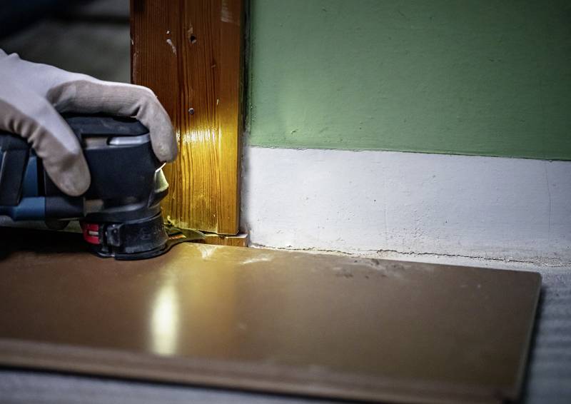 Eine Person benutzt ein Elektrowerkzeug, um die Basis eines hölzernen Türrahmens zu schneiden, wobei eine grüne Wand und ein geneigtes Holzbrett in der Nähe sind.