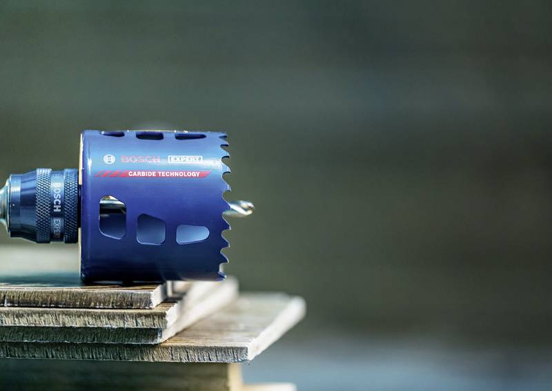 Eine blaue Hartmetall-Lochsäge mit dem Logo der Marke ist an einem Bohrer befestigt, der auf einem Stapel von Holzbrettern ruht.