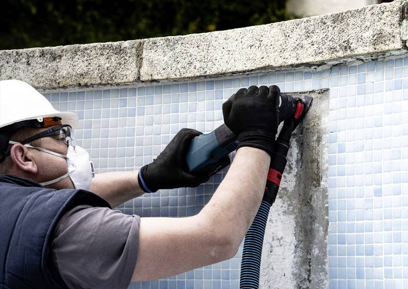 Ein Arbeiter mit Schutzhelm und Maske nutzt ein Elektrowerkzeug, um die Kante eines Swimmingpools zu reparieren, der mit blauen Mosaikfliesen ausgelegt ist.