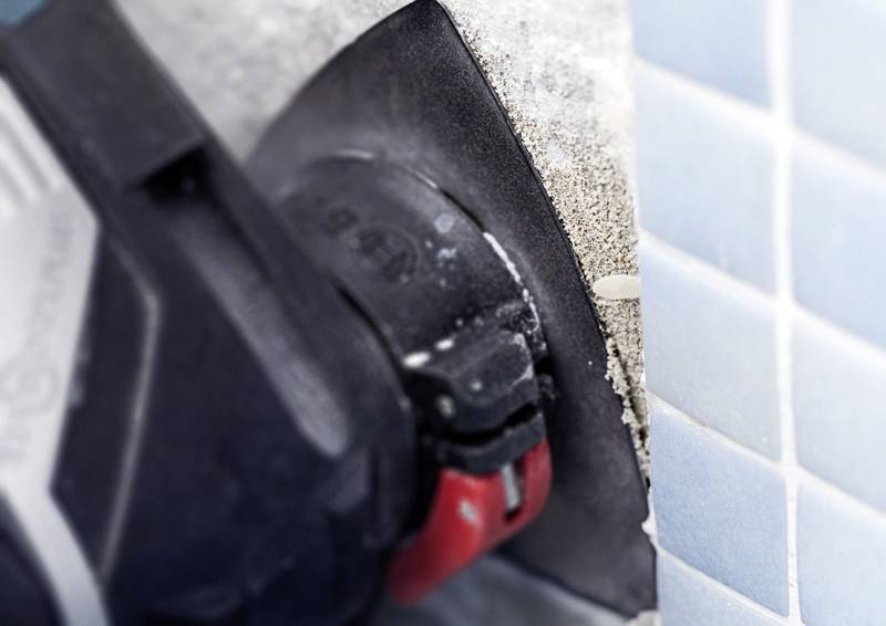 Ein Nahaufnahme eines Elektrowerkzeugs, das durch eine Wand oder Oberfläche schneidet, mit blauen Kacheln auf der rechten Seite, das Staub und Trümmer des Schnitts zeigend.