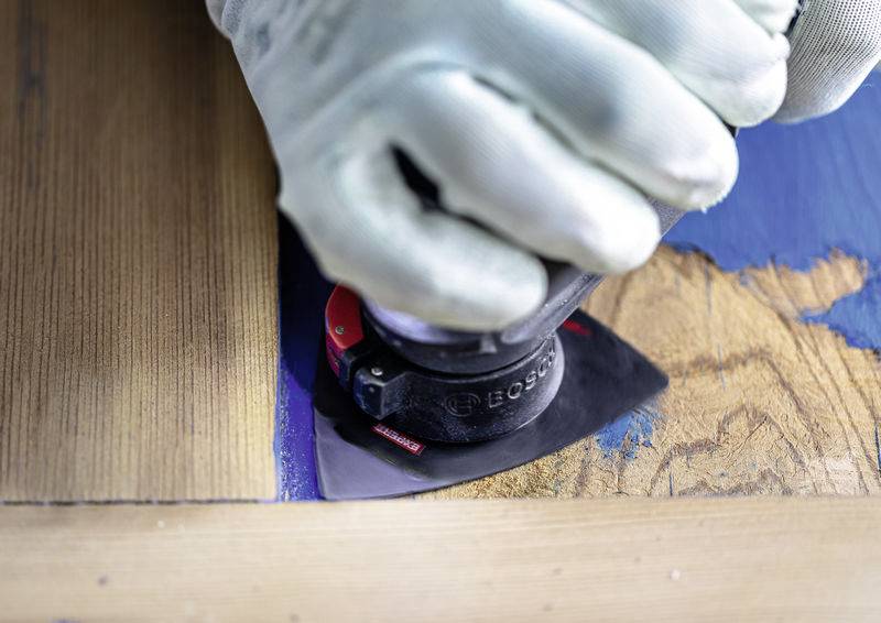 Eine Person, die ein Bosch-Multifunktionswerkzeug mit Oszillation verwendet, um blaue Farbe von Holz abzuschleifen, mit Fokus auf das Schleifpad des Werkzeugs in Aktion.