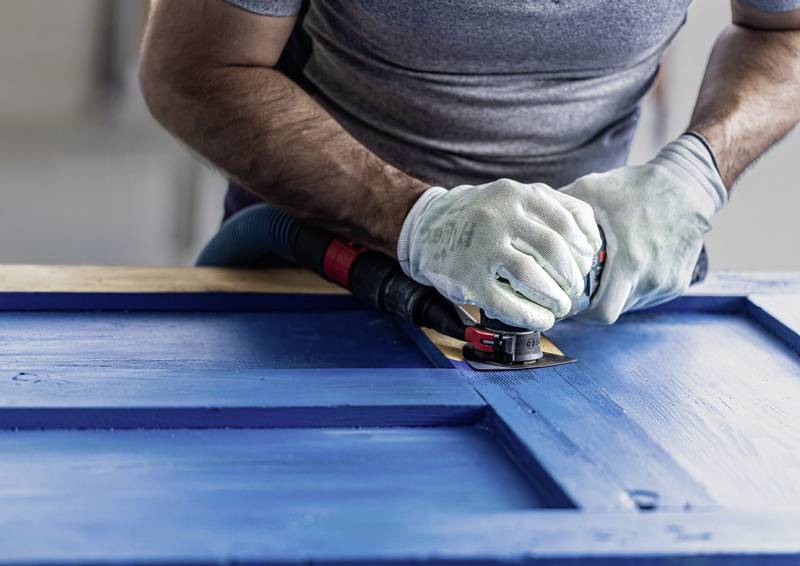 Person mit Handschuhen, die eine blaue Holztür mit einem Elektrowerkzeug abschleifen, wobei der Fokus auf dem Glätten der Oberfläche liegt.